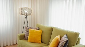 Un sofà verd amb dos coixins grocs i almoades estampades, col·locat davant d'una làmpada de peu i cortines blanques.