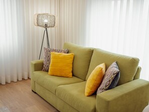 Ein grünes Sofa mit zwei gelben Kissen und gemusterten Zierkissen, vor einem Stehlampen und weißen Vorhängen platziert.