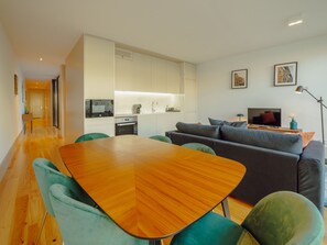 Um espaço de estar moderno com mesa de jantar, cadeiras verdes, sofá cinza, televisão e área de cozinha.