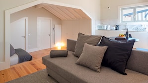 Ein modernes Wohnzimmer mit einem grauen Sofa, einem kleinen Hocker und Holzboden. Es gibt eine Küchenzeile mit Fenster und Mikrowelle.