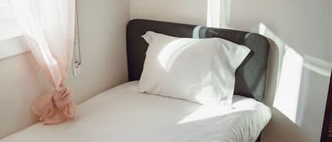 A single bed with white bedding, a dark headboard, and a window with sheer curtains.