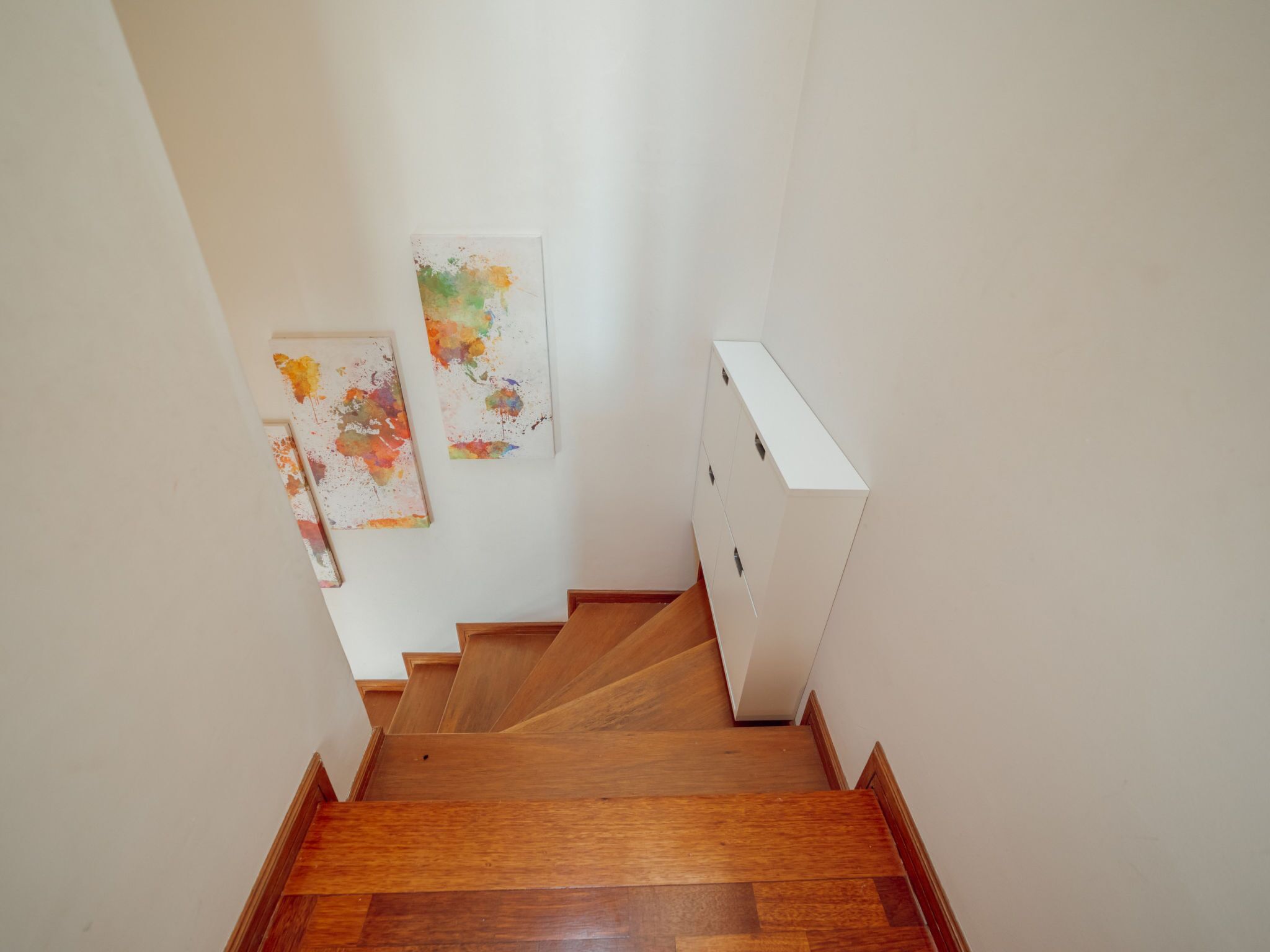 Een houten trap die uitkomt op een vloer met witte muren en twee abstracte schilderijen.
