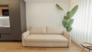 Una sala moderna con un sofá color beige, piso de madera y una cocina al fondo.