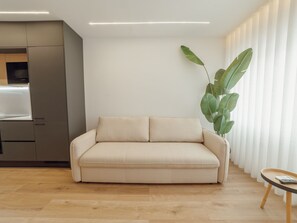Ein modernes Wohnzimmer mit einem beigefarbenen Sofa, Holzboden und einer Küche im Hintergrund.