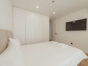 Ein modernes Schlafzimmer mit einem großen Bett, einem Kleiderschrank, einem Flachbildfernseher und einer Tür zu einem anderen Raum.