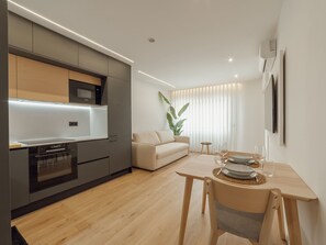 Eine moderne Küche mit integrierten Geräten, ein Essbereich mit Tisch für zwei Personen und ein Wohnbereich mit Sofa und einer großen Pflanze.