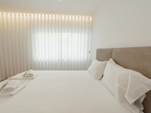 Una habitación de hotel con cama, ropa de cama blanca, cabecero y ventana con cortinas.