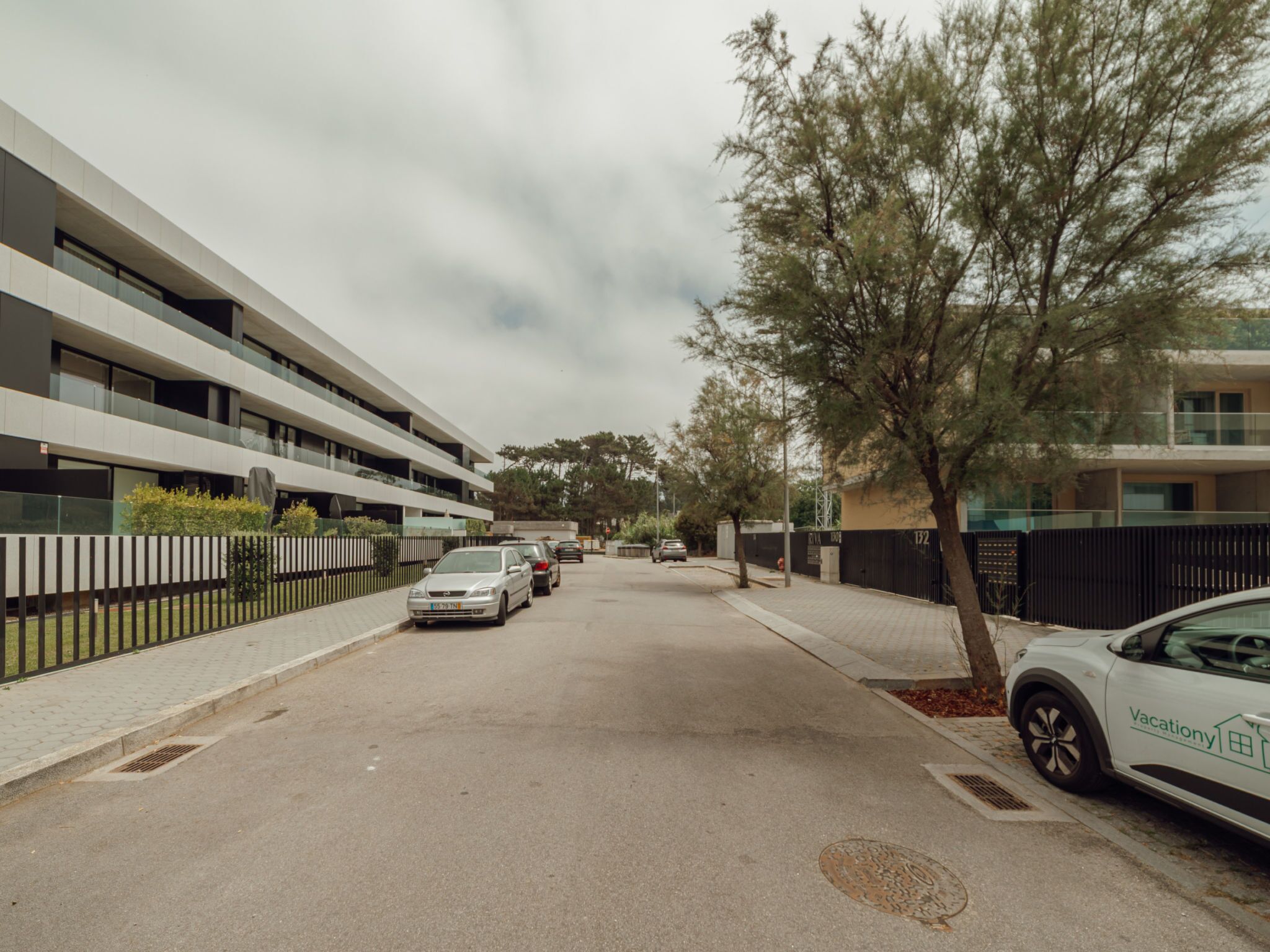 Una calle residencial con edificios modernos, coches estacionados y una acera arbolada.