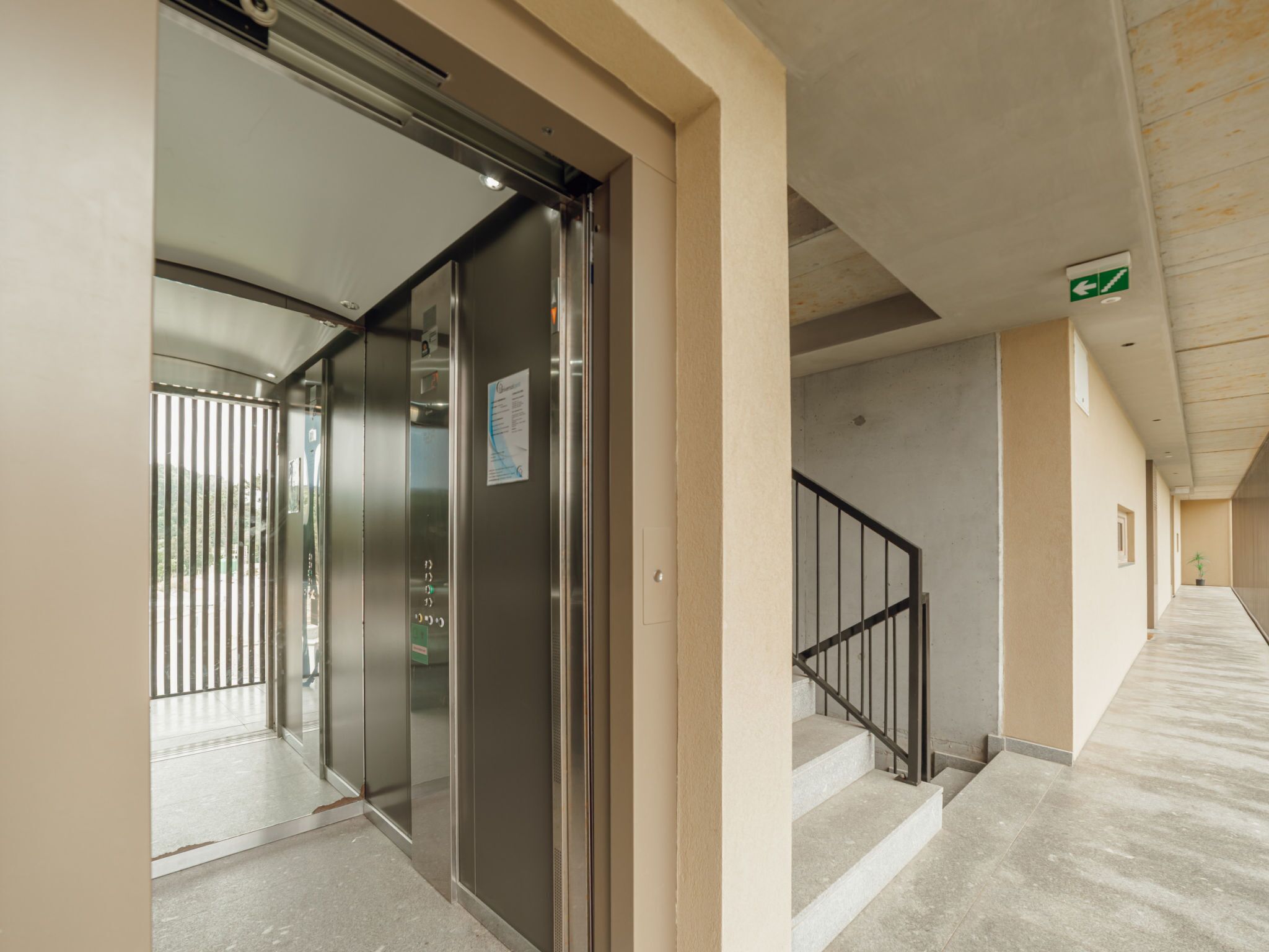 Un corridoio interno con porta a vetro per ascensore, una scala con ringhiera in metallo e un corridoio con indicazione a soffitto.