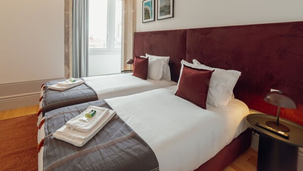 A hotel room with two beds, a window with curtains, a bedside table with a lamp, and artwork on the wall. - Apartment  by Interhome (Porto)