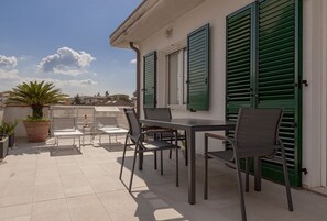 Una terraza con una mesa y sillas, plantas en macetas y un edificio con contraventanas verdes.