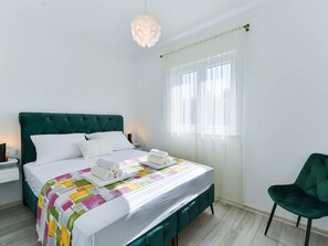 Habitació d'hotel amb un llit, una cadira tapissada de verd, una taula de nit i una finestra amb cortines primes.