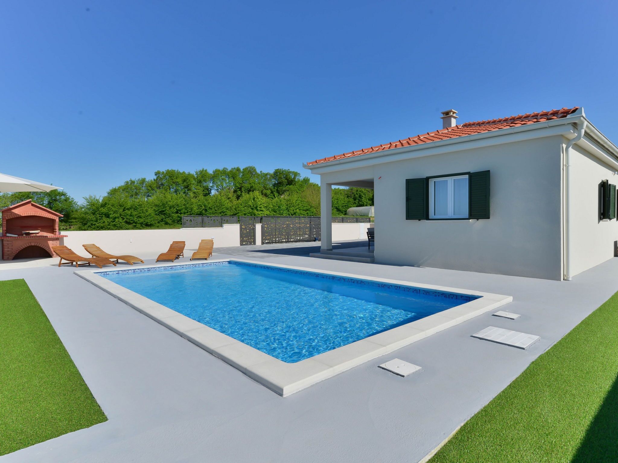 A rectangular swimming pool with a white deck, surrounded by green artificial grass. There are wooden lounge chairs and a small brick structure, possibly a sauna or hot tub, to the left.