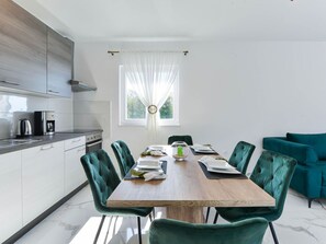 Eine moderne Küche mit Essbereich, inklusive einem Holztisch, grünen Polsterstühlen und Blick ins Grüne durch ein Fenster.