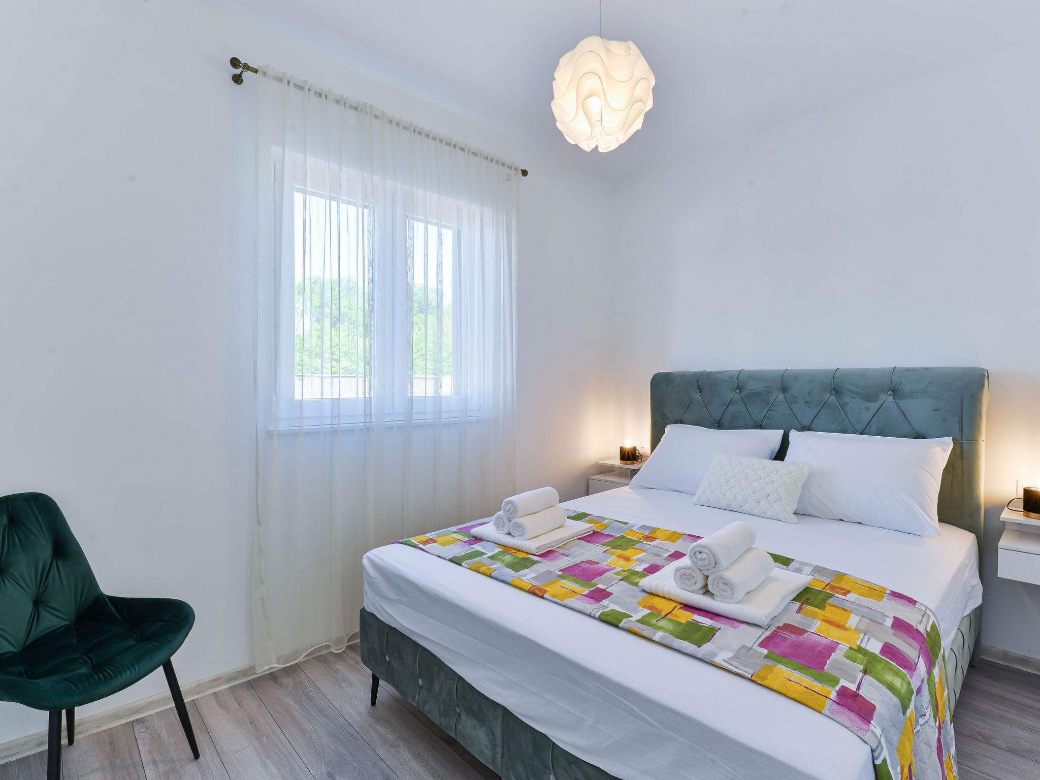 A hotel room with a bed, a green chair, a window with sheer curtains, and a hanging light fixture.