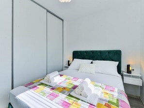 Ein ordentlich bezogenes Bett mit einem farbenfrohen Quilz, weißen Kissen und aufgerollten Handtüchern. An beiden Seiten des Bettes stehen Nachttische mit Lampen.