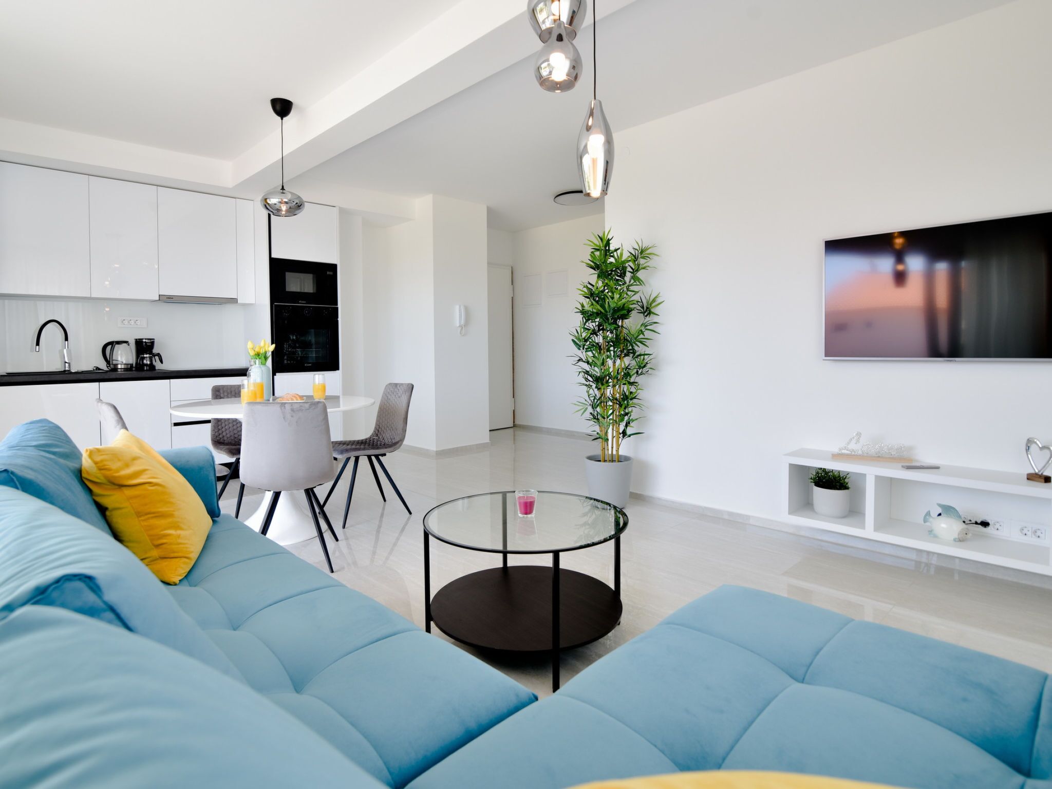 A modern living room with a blue sofa, a glass coffee table, and a dining area with chairs and a kitchen in the background.