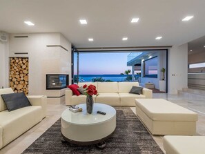 A modern living room with a large window overlooking the sea, a fireplace, and a round coffee table with a vase of flowers. - Luxury Villa Michele with infinity pool by Interhome (Rovanjska)