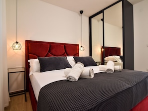 Ein ordentlich bezogenes Bett mit Kissen und Handtüchern, ein rotes, mit Stoff überzogenes Kopfteil und ein großer Spiegel an der Wand.