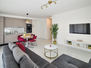 Ein modernes Wohnzimmer mit einem grauen Sofa, roten Sesseln, einem kleinen weißen Tisch und einem an der Wand befestigten Fernseher.