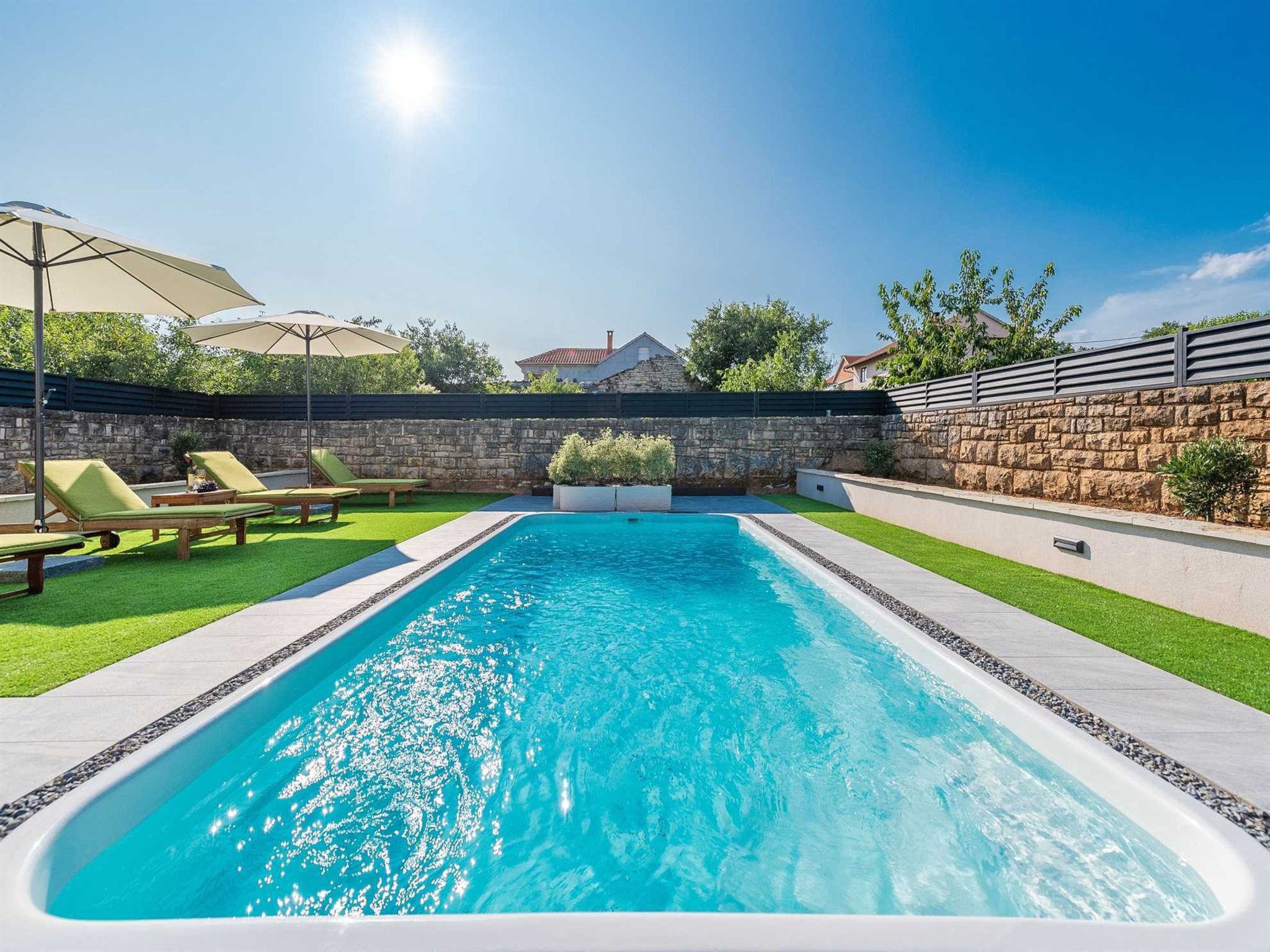 Una piscina rectangular amb una paret de pedra i una zona verda amb gandules i ombrelles.