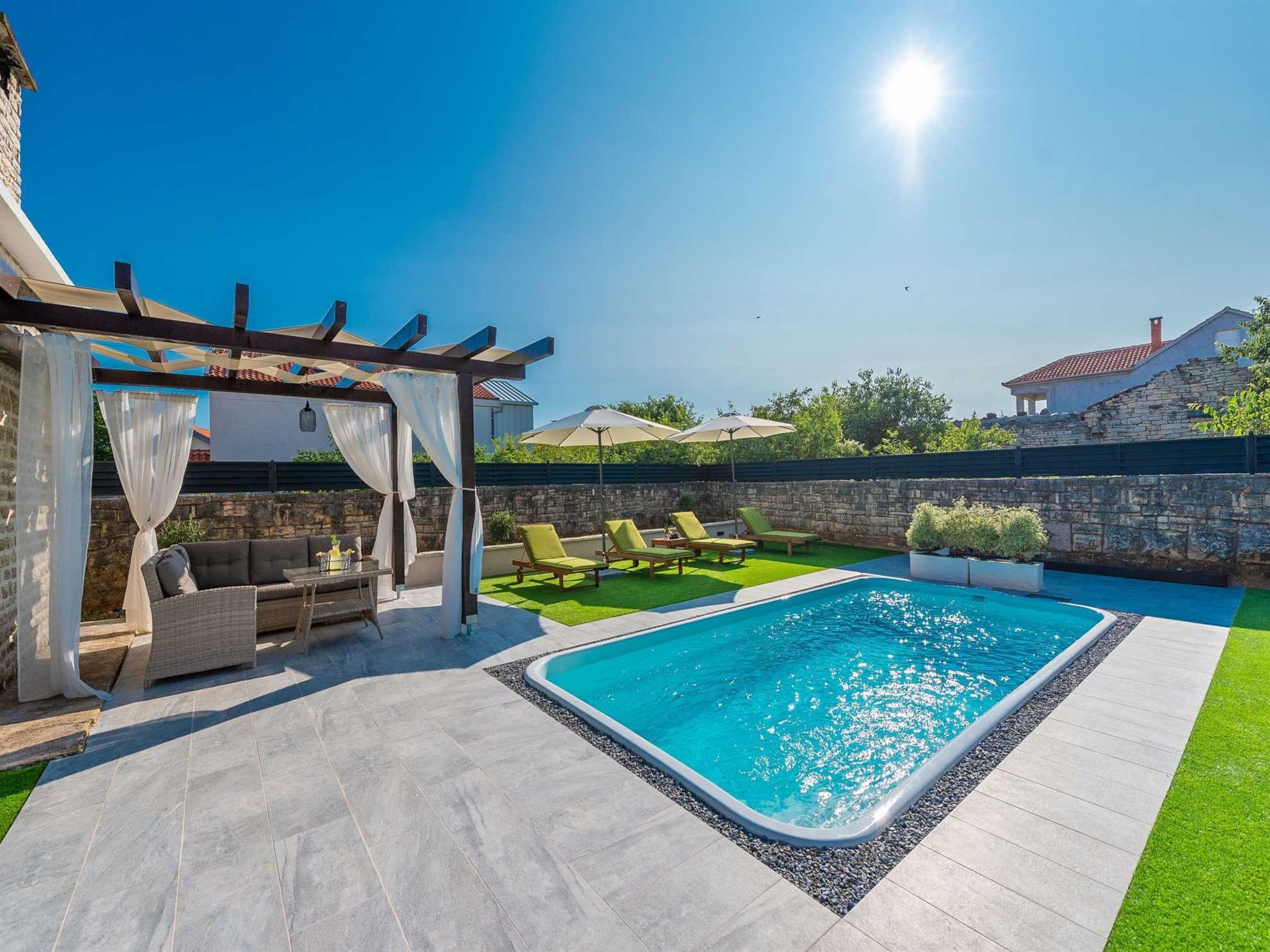 A swimming pool with lounge chairs, a pergola with white curtains, and a stone wall.