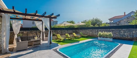 Ein Schwimmbecken mit Liegestühlen, eine Pergola mit weißen Vorhängen und eine Steinmauer.