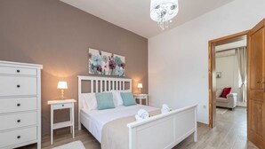 A bedroom with a bed, bedside tables, a dresser, a chandelier, and a painting on the wall.
