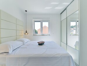 A bedroom with a bed, a bedside table, a hanging light, and a window with a view of buildings. - Apartment  by Interhome (Zadar/Bibinje)