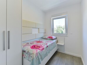 Quarto pequeno com cama de solteiro, janela, mesa de cabeceira e guarda-roupa.