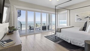 Ein Schlafzimmer mit einem großen Bett, einem Schreibtisch mit einem Buch, einem Stuhl und einem Balkon mit Meerblick.