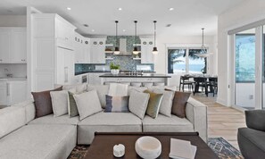 A modern kitchen with white cabinets, a central island, pendant lighting, and a view to the outside. - Sea Swept | Luxury Beachfront Estate (Bradenton Beach)