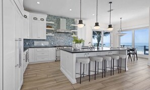 A modern kitchen with white cabinetry, a large island with bar stools, and a view of the ocean. - Sea Swept | Luxury Beachfront Estate (Bradenton Beach)