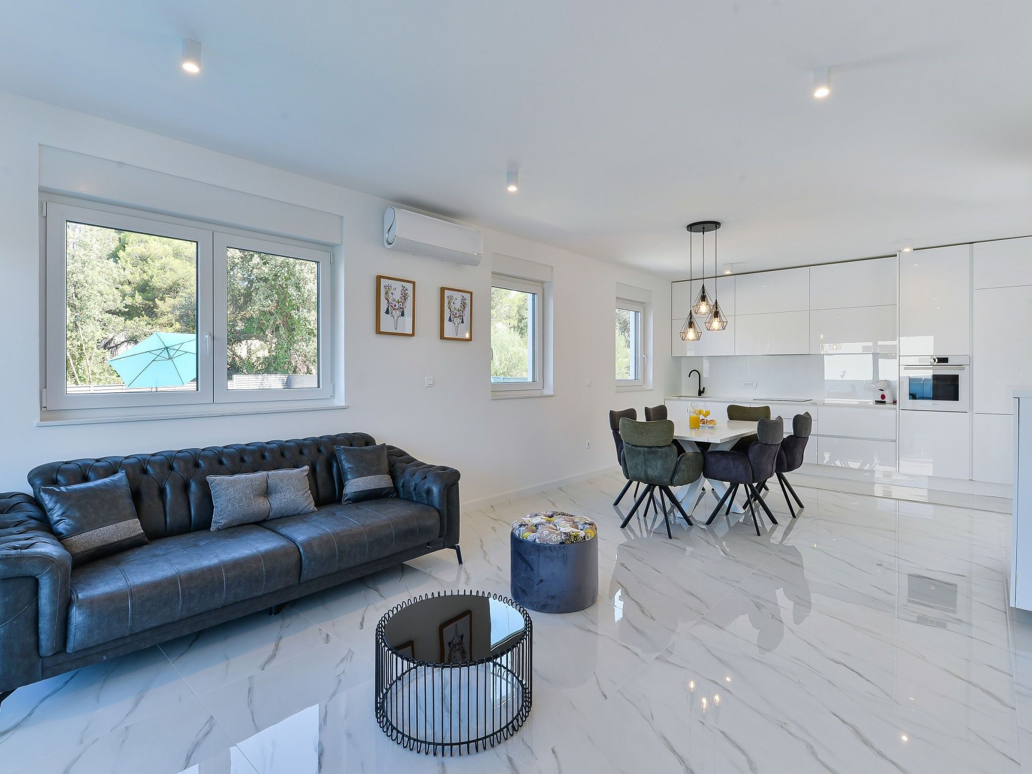 Una sala de estar moderna con sofá, mesa de comedor y sillas, comunicada con una cocina con armarios blancos y una lámpara colgante.