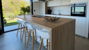 Eine moderne Küche mit einer zentralen Kochinsel, Barhocker, einer Kaffeemaschine und Blick ins Freie.
