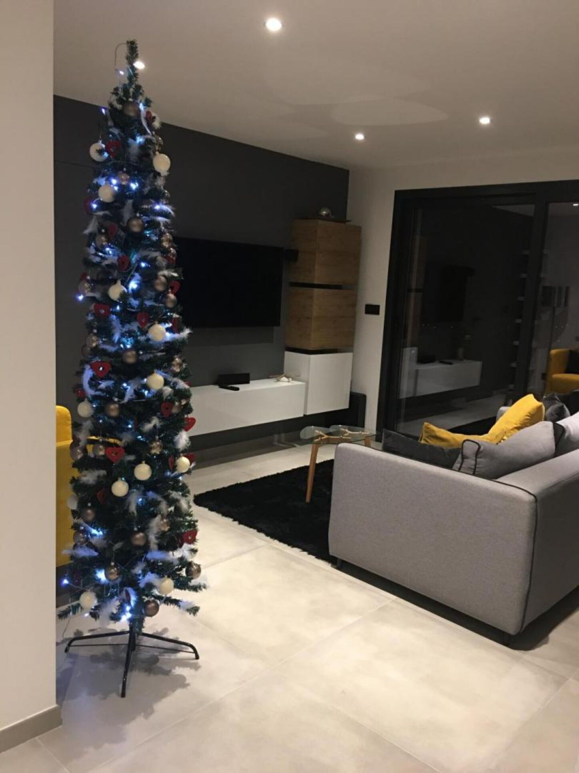Ein modernes Wohnzimmer mit einem hohen Weihnachtsbaum, einem grauen Sofa und einer Holzkassette.