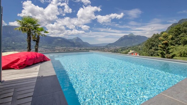 Ein klares, blaues Schwimmbecken mit einer Holzterrasse, einem roten Kissen und Blick auf Berge und eine Stadt im Hintergrund.