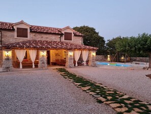 Ein zweigeschossiges Steinhaus mit einem Ziegeldach, einer Schotterauffahrt und einem Pool im Hintergrund.