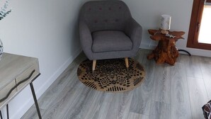 Una habitació amb una poltrona gris, un tapet rodó, una taula de fusta i un tamboret de fusta singular.
