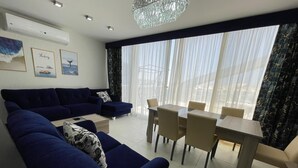 Ein modernes Wohnzimmer mit einer Sofagarnitur, einem Esstisch mit Stühlen und einem großen Fenster mit transparenten Vorhängen.
