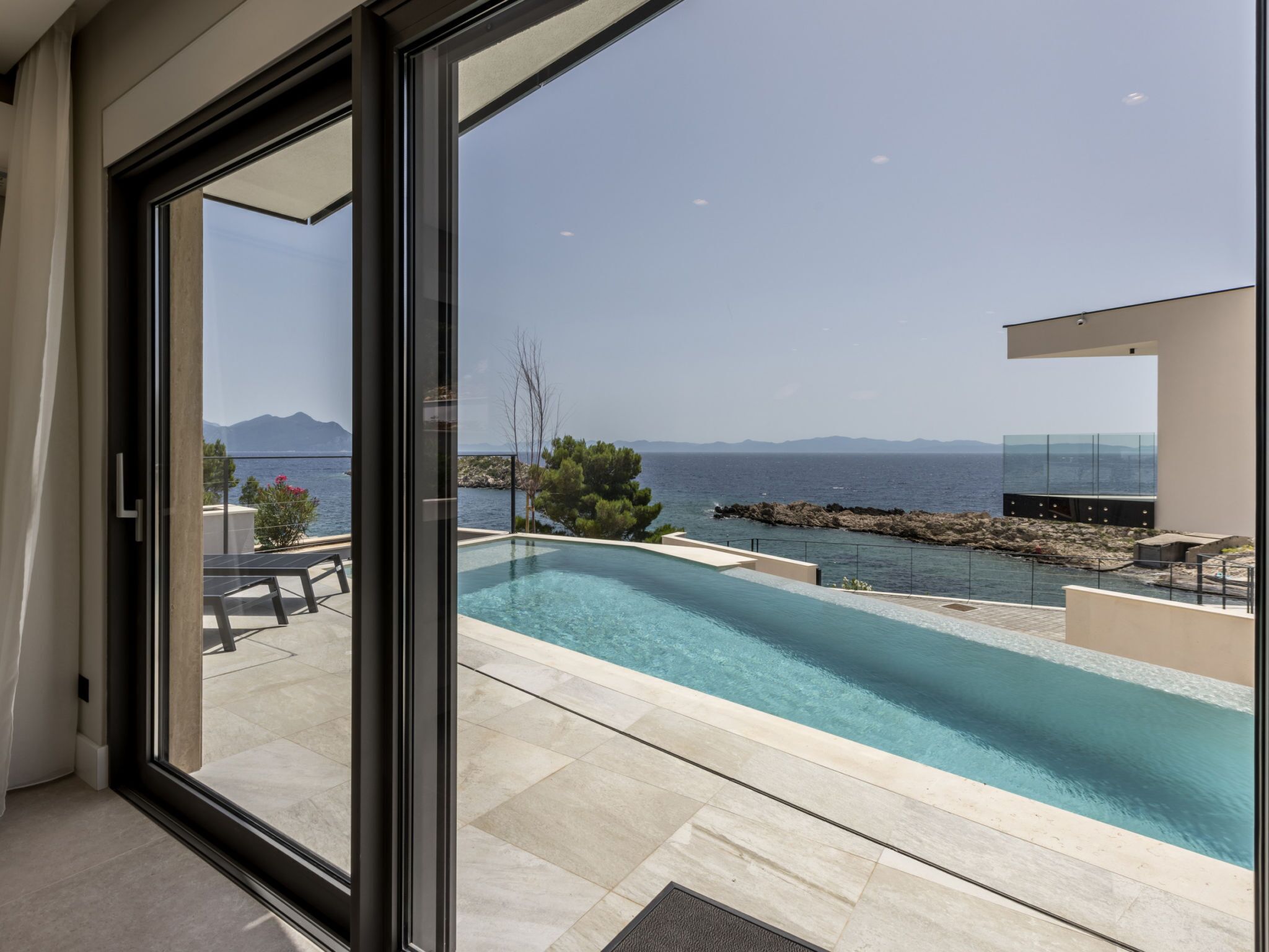 A modern swimming pool with clear blue water, surrounded by a stone deck, and overlooking a calm sea with distant mountains.