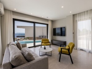 A modern living room with a sofa, two armchairs, a coffee table, and a flat-screen TV. There is a large sliding glass door leading to a balcony with a pool view. - Beachfront villa Luce with seaview and pool by Interhome (Trstenik/Dingač)