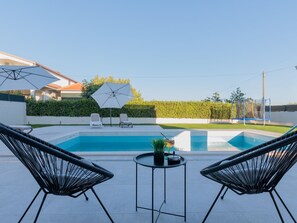 Ein Schwimmbecken mit zwei Liegestühlen und einem Tisch, umgeben von einer Hecke und blauem Himmel.