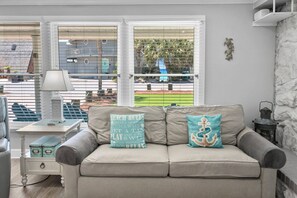 Una sala de estar con un sofá, dos almohadones decorativos, una mesita con una lámpara y vistas amplias a través de grandes ventanales.