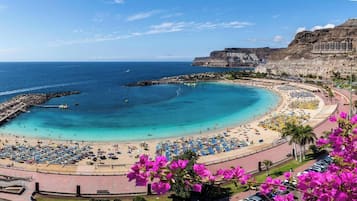 Une plage côtière avec un rivage incurvé, une promenade bordée de parasols et une falaise sur laquelle se trouvent des bâtiments.