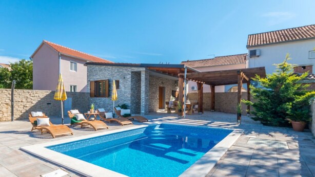 Ein Swimmingpool mit Liegestühlen und Sonnenschirmen, umgeben von Steinmauern und einer überdachten Terrassenanlage.