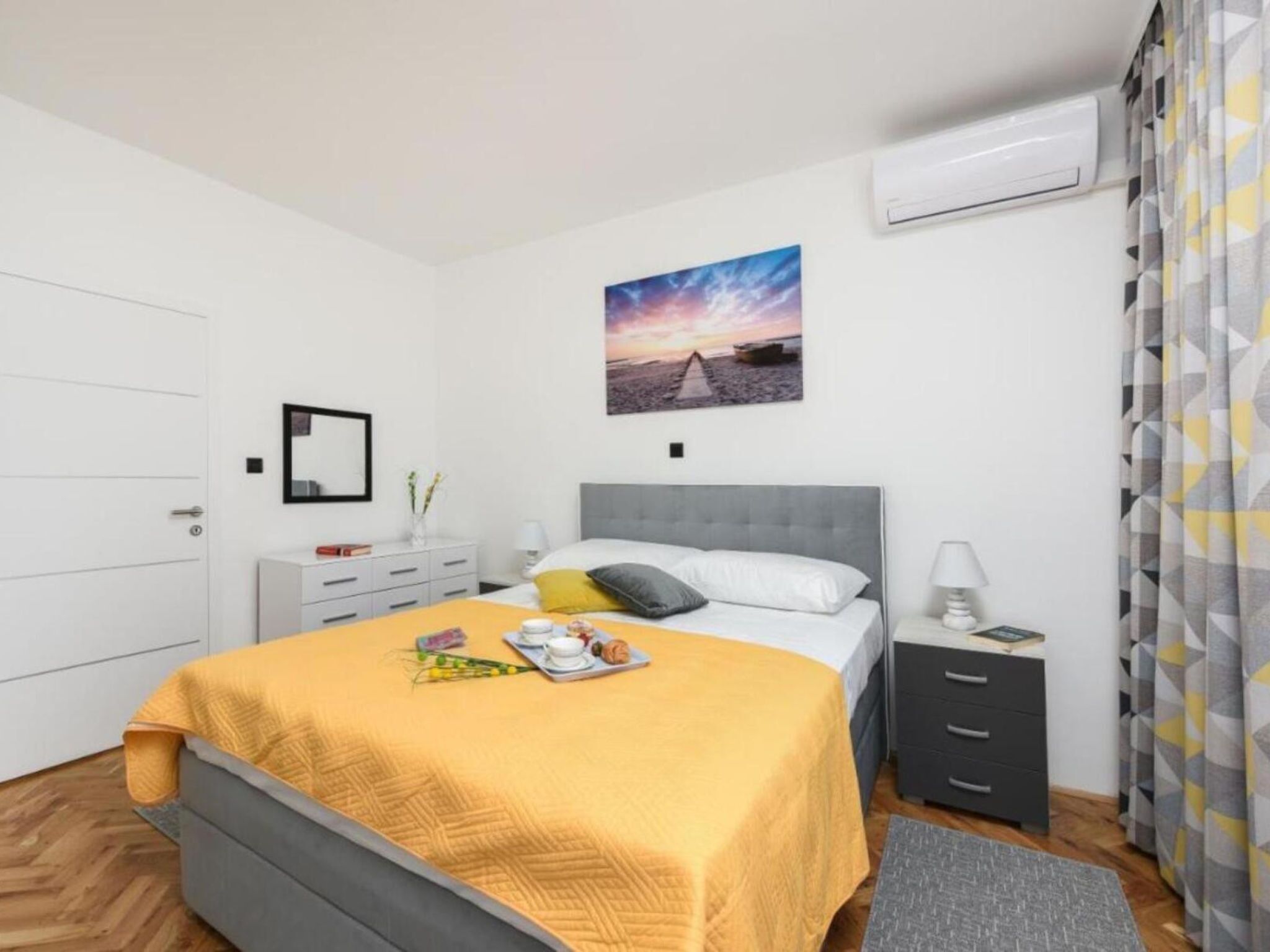 A hotel room with a bed, bedside table, a nightstand, a picture on the wall, and an air conditioning unit.