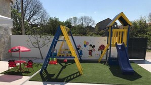 Ein Spielplatz mit Rutsche und Klettergerüsten, umgeben von einer gepflasterten Fläche mit einem roten Sonnenschirm und Sitzgelegenheiten.