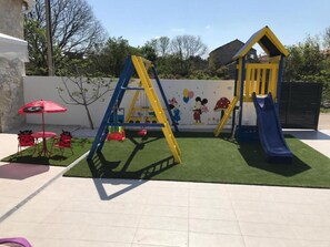 A playground with a slide and climbing structures, surrounded by a paved area with a red umbrella and seating. - Villa Mare with heated pool and gym by Interhome (Crno)