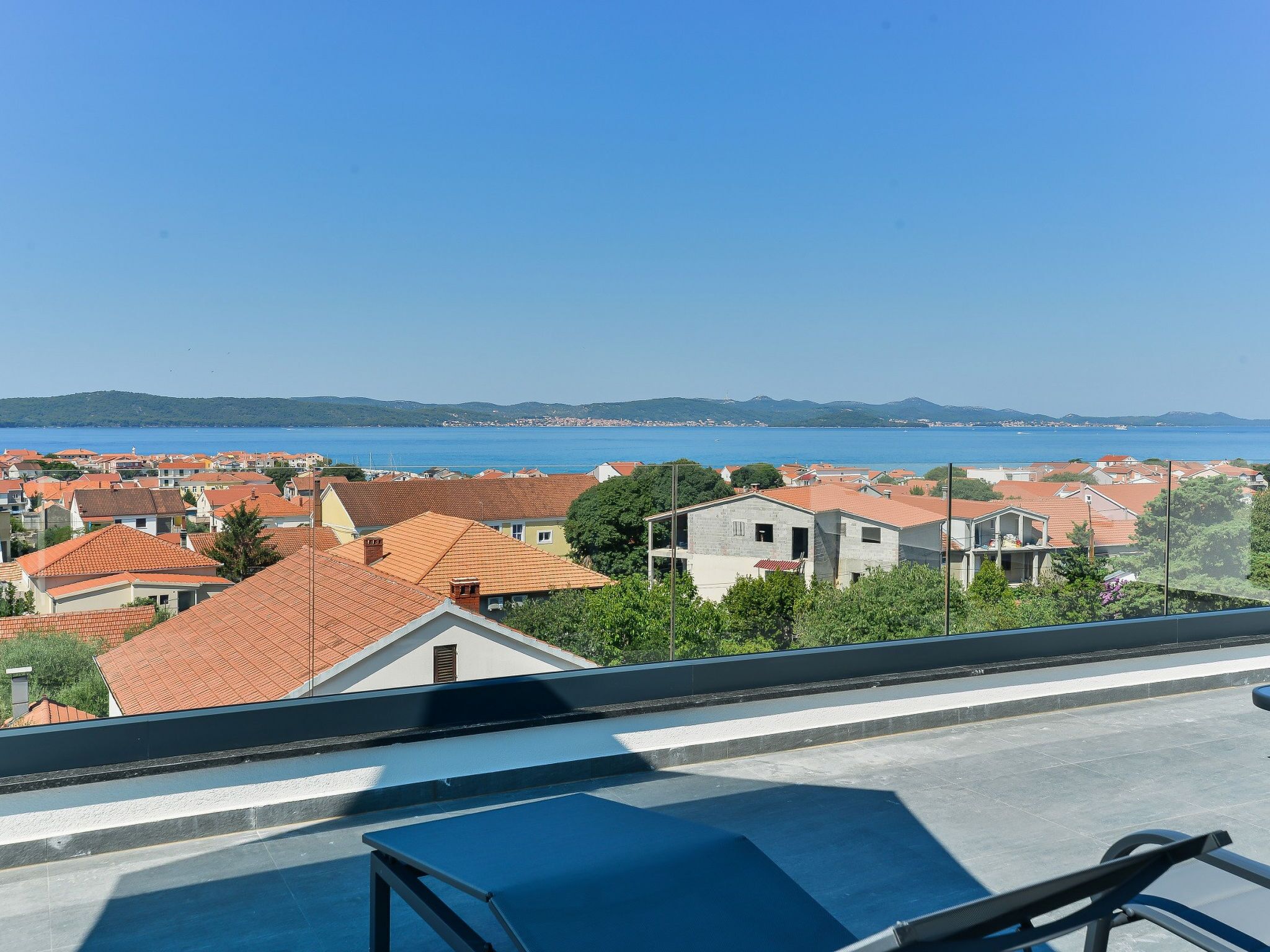 Zona residencial com telhados de telha vermelha, céu azul límpido e vista para o mar e colinas ao longe.